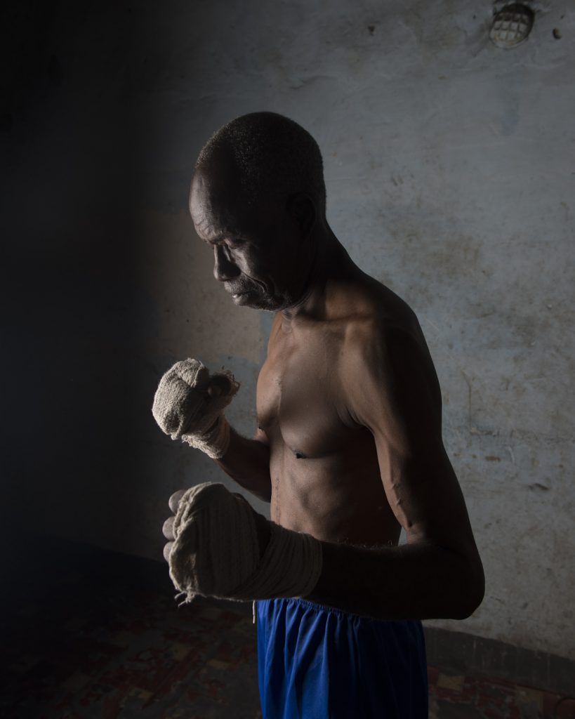 Kinshasa, République démocratique du Congo. César Sinda, ex-champion de boxe. Né en 1948, d’une famille de douze enfants, César devient boxeur après que sa sœur se fasse attaquer à l’école. Il devient champion national des poids légers, puis est couronné champion d’Afrique en Zambie. A son retour à Kinshasa, Mobutu est absent, mais laisse des ordres pour lui attribuer une maison et une voiture neuve, dont il ne verra jamais la couleur. En 1969, il est envoyé aux jeux olympiques de Mexico. Quand Mohamed Ali arrive à Kinshasa, César Sinda est au Etats-Unis, dans l’espoir d’y suivre un entrainement professionnel pour devenir champion du monde des poids légers. Parti avec six dollars en poches, un pasteur de New York le prend sous son aile et l’adopte officiellement. Après plusieurs défaites, sa carrière décolle enfin. Mais en 1978, il retourne au Congo pour enterrer son père. Quelques mois plus tard, sa sœur décède, et son passeport est volé. Il décide alors de rester à Kinshasa, et devient entraineur. Il sera couronné de l’ordre du Léopard par Mobutu.Interview réalisée le 21/11/2015.Portrait posé à son domicile.